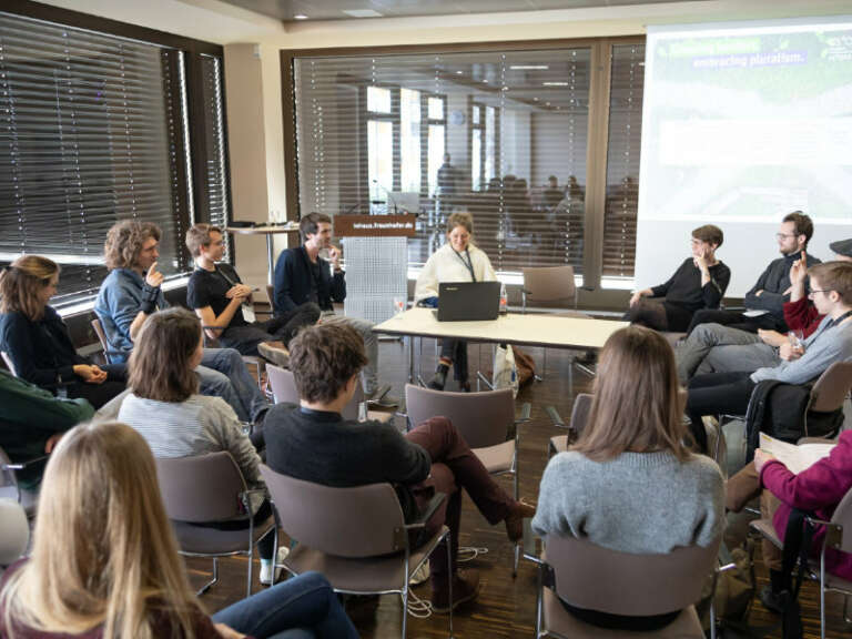 Studierende sitzen in einem Stuhlkreis im Seminarraum, eine Frau hält einen Vortrag.